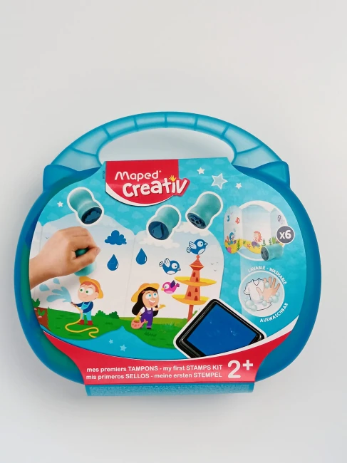niebieska-walizeczka-z-tekturową-owijką-z-napisem-Maped-Creative-Mój-pierwszy-zestaw-do-stemplowania-2+-widać-uśmiechnięte-dzieci-które-biegają-po-podwórku-i-fontannę-z-ptaszkami.jpg